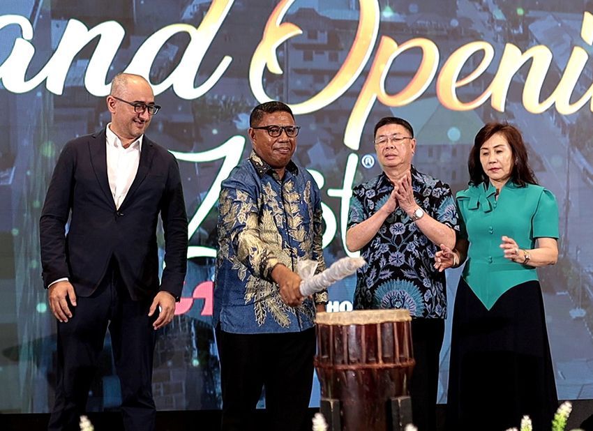 Four people at a grand opening event. Man in patterned shirt strikes a drum with a mallet.