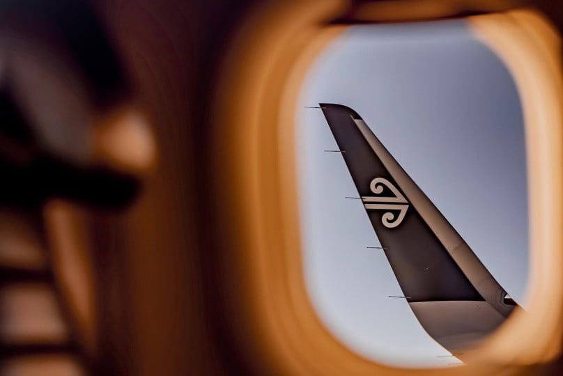 Woman smiles while holding a phone attached to a tripod outdoors. She wears a white blazer and jeans. Red brick building in background.