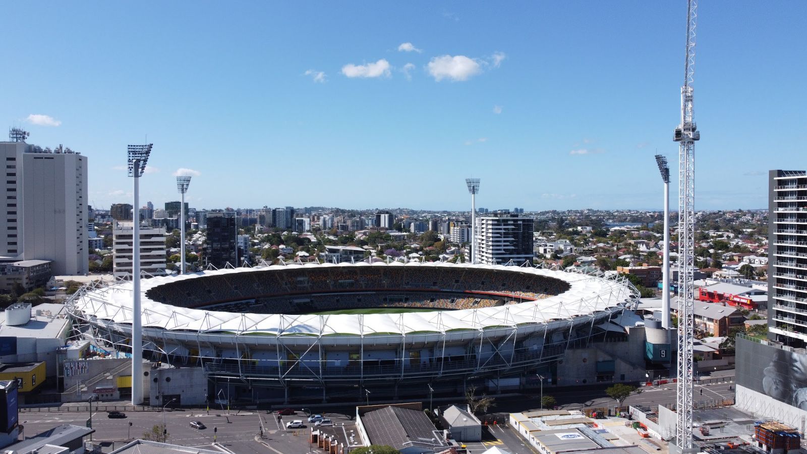 Aerial view of a stadium with a playing field, surrounded by seating, tall light towers, and a cityscape on a sunny day.