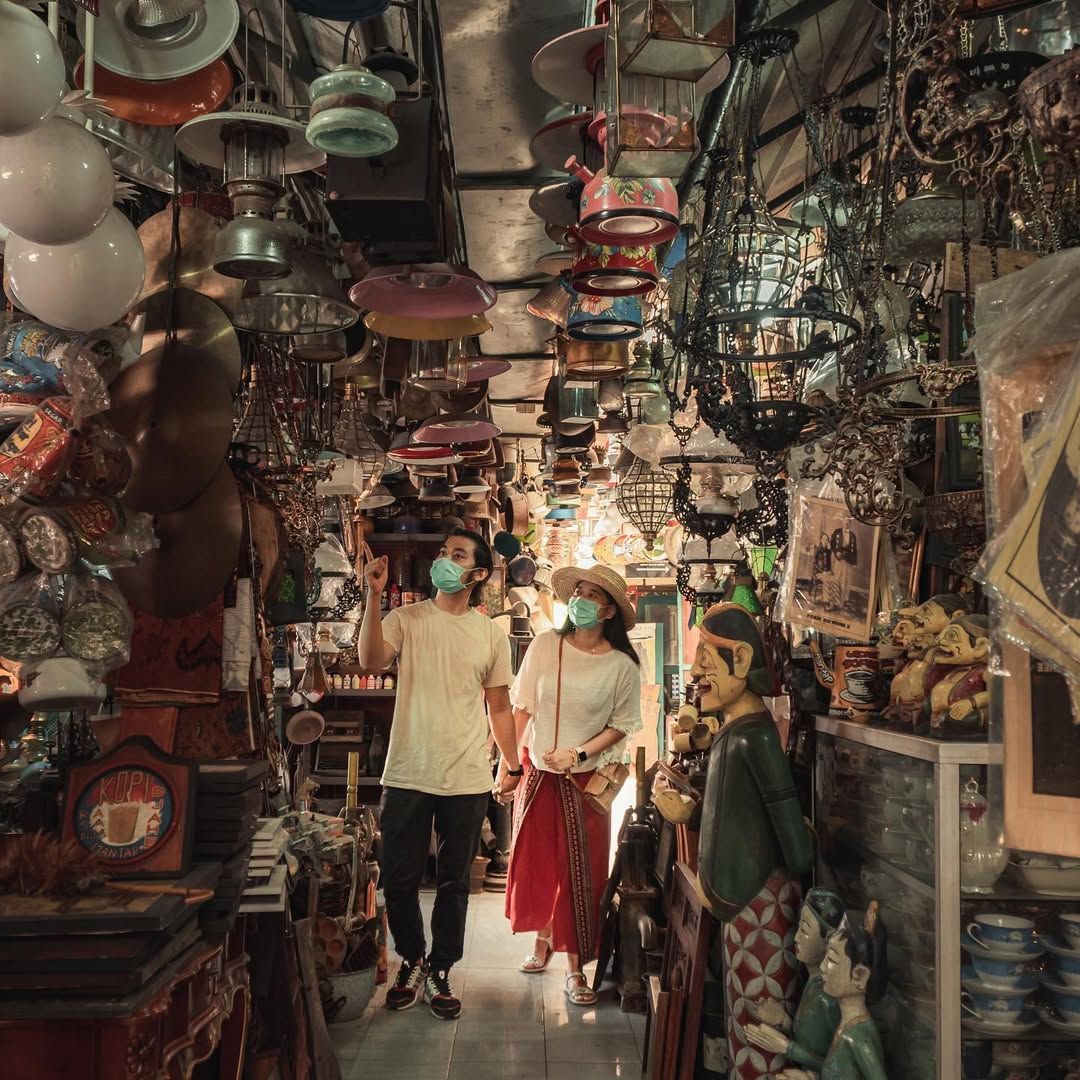 Couple wearing masks walks through a crowded antique shop with hanging lamps.