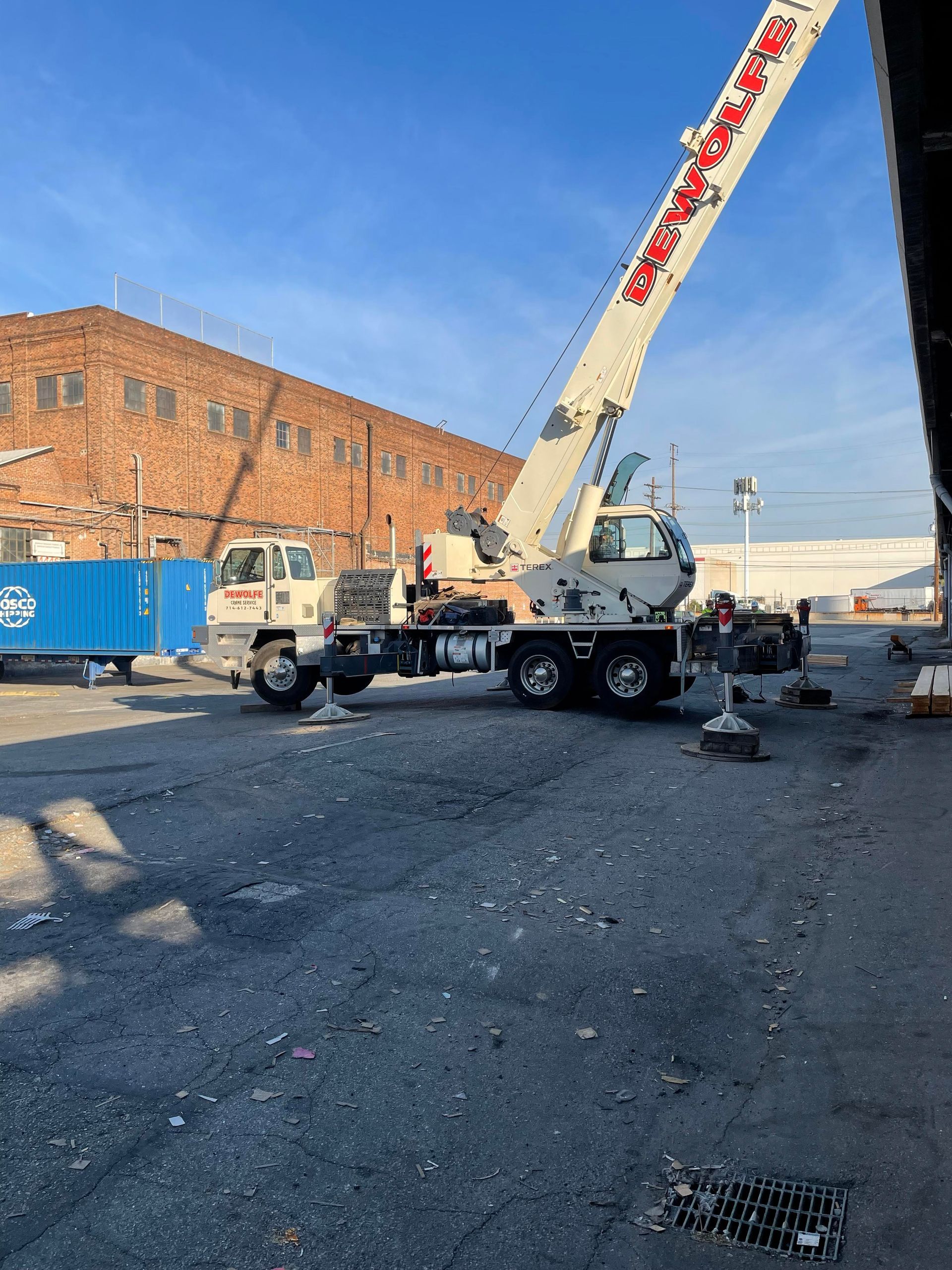 Crane on a concrete surface, boom raised, near a brick building and a blue container.