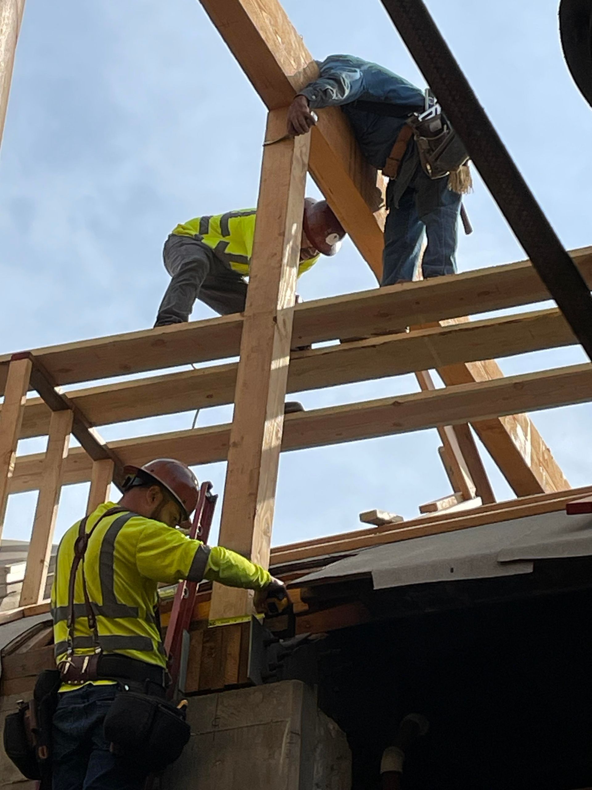 Construction workers on a roof, building a wooden structure. One in a harness, two others with lumber, against a blue sky.