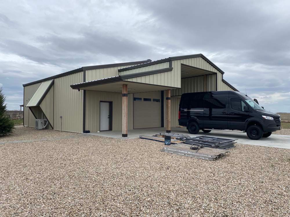 Tan metal building with black van parked in front, gravel driveway, cloudy sky.