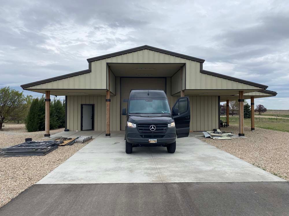 Black van parked in front of a tan barn-style building with open door; cloudy sky.