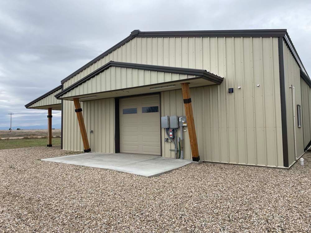 Tan metal building with a garage door and concrete pad, supported by angled wood beams.