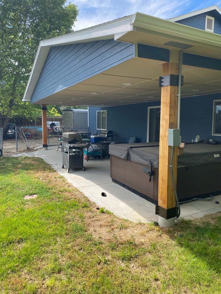 A covered patio with blue siding, supported by wooden posts. Contains a grill and a hot tub.