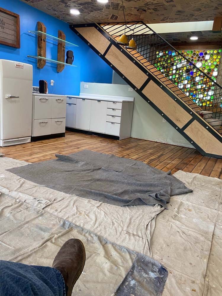 Person's feet with work boots, floor and wall painting in progress; kitchen/stairwell.