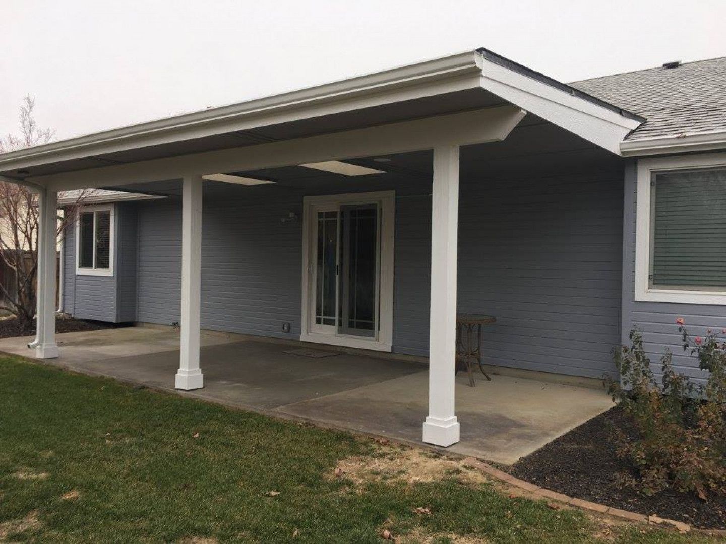 Covered patio with white columns, concrete flooring, and sliding glass door. Gray house with a green lawn.