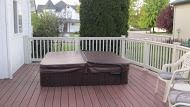 Hot tub with cover on a wooden deck surrounded by a white railing, with a house and trees in the background.