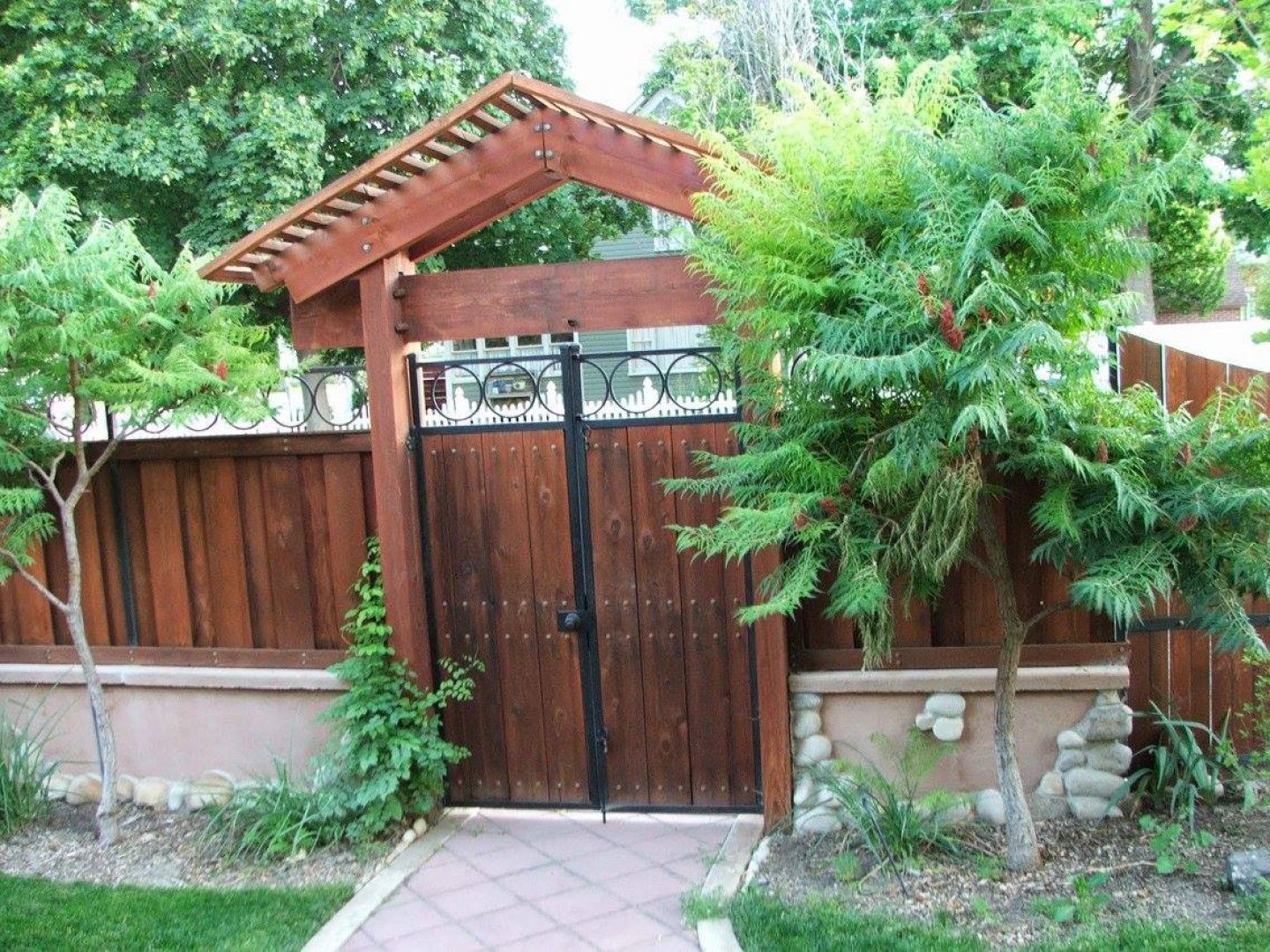 Wooden gate with pergola roof, leading to a path, flanked by trees and a decorative fence.