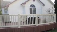 Beige deck with white railing in front of a white house with a round window.