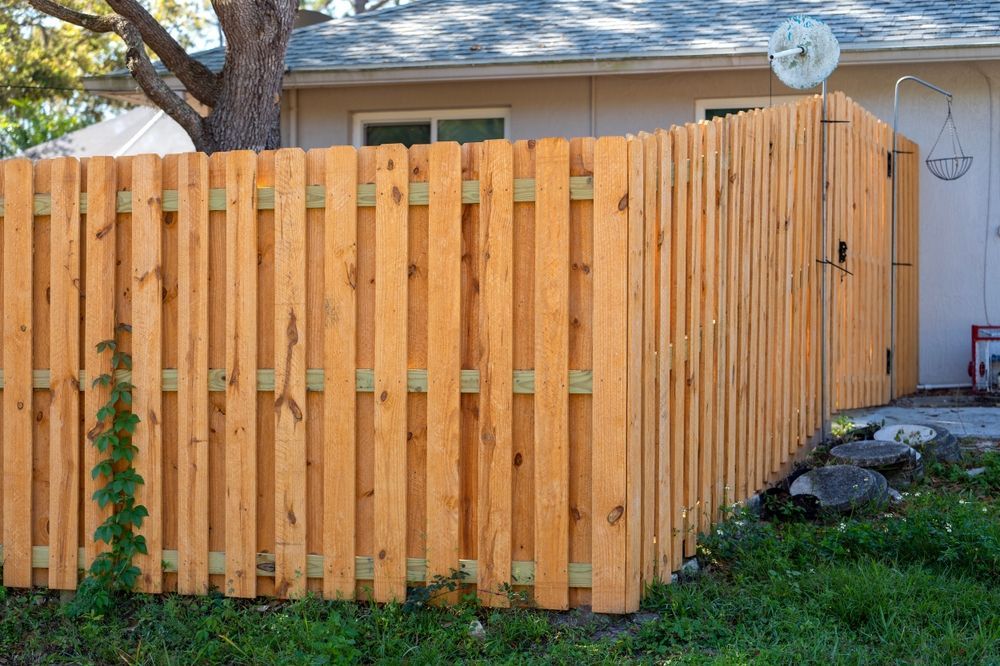 Wooden privacy fence in a backyard, partially blocking a light-colored house. Green grass in foreground.