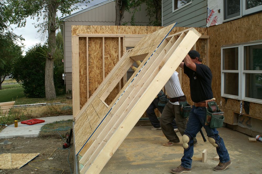 Construction workers lift a wall frame to attach it to an existing house; outdoors.