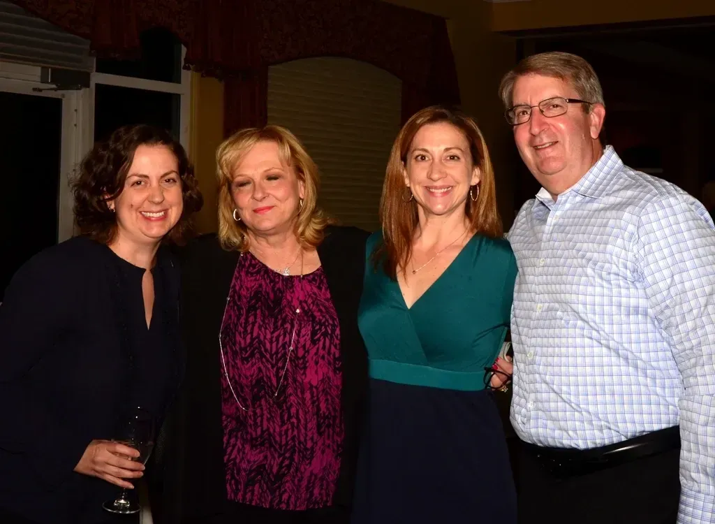 Four people smiling, posing together indoors. A woman in a teal dress stands next to a man in a blue button-down shirt.