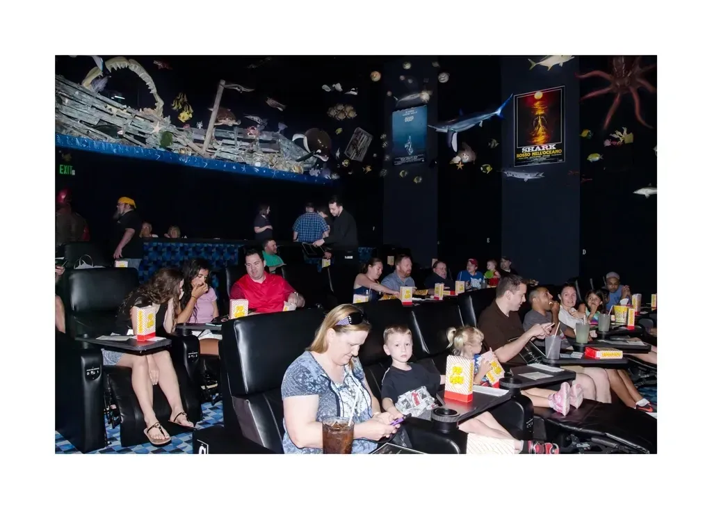 People watch a movie in a dark theater with themed decorations. Seats have trays with snacks.