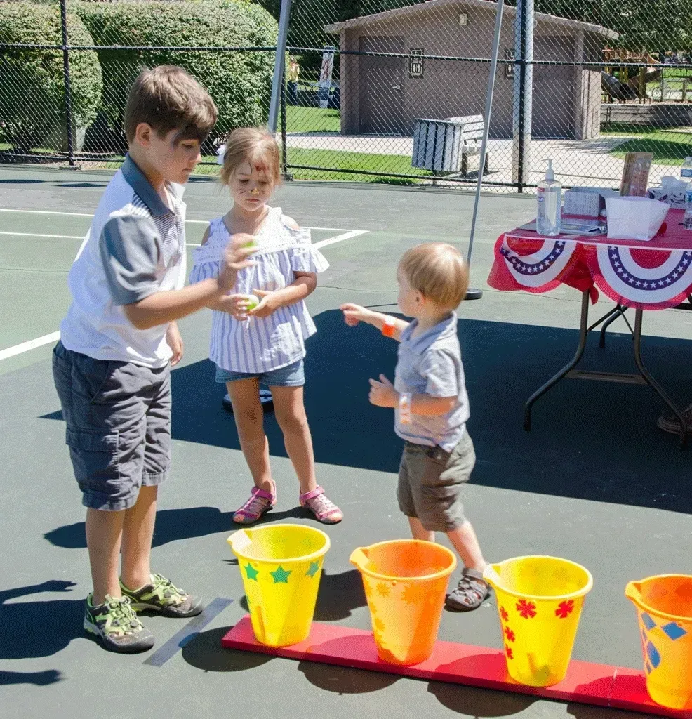 Children playing a ball toss game with colorful buckets on a tennis court.