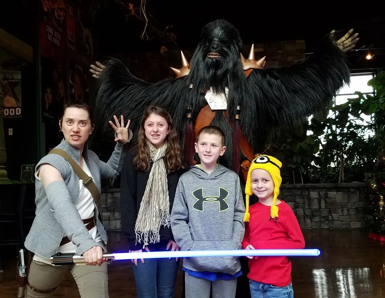 People pose with a furry figure and lightsaber. One person dressed as Rey, others smiling. Inside a building.