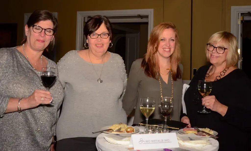 Four women at an event holding wine glasses, smiling.  Table with snacks in front of them.