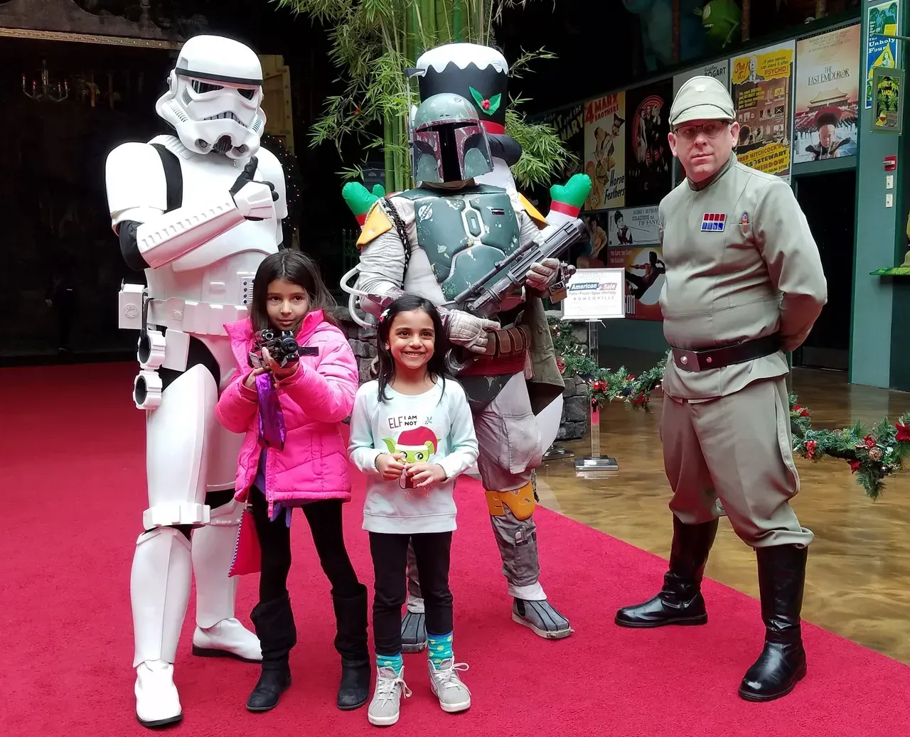 Two girls pose with a Stormtrooper, Boba Fett, and an Imperial officer on a red carpet.