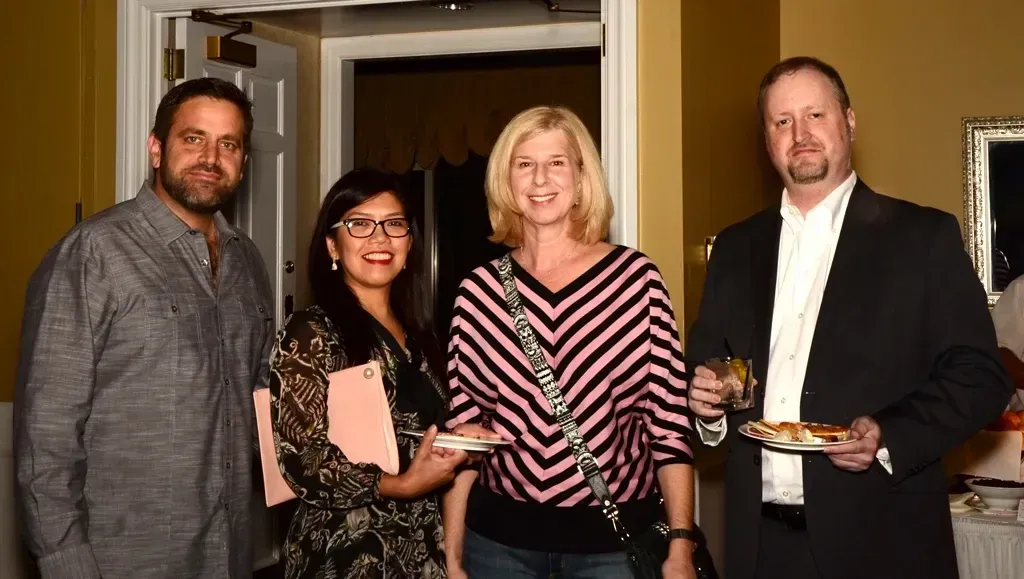 Four people at an event, holding plates of food. Inside a building, well lit, smiling.