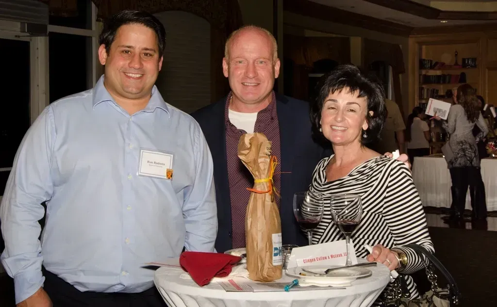 Three people at an event, smiling near a table with a wine bottle, woman holding a wine glass.