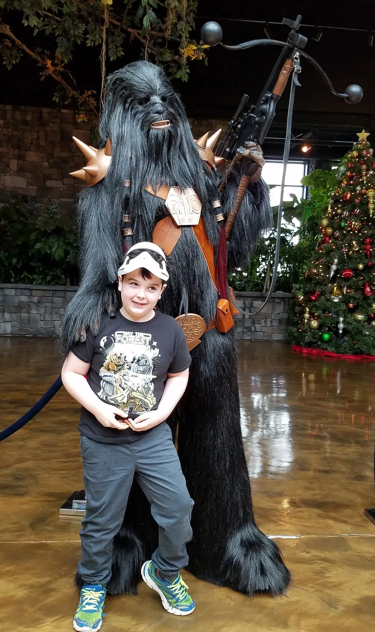 A child smiles, poses next to a tall, furry Chewbacca statue with a crossbow in a room, Christmas tree in the background.