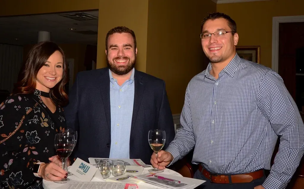 Three people at an event, holding wine glasses, smiling. A table with brochures is in front.