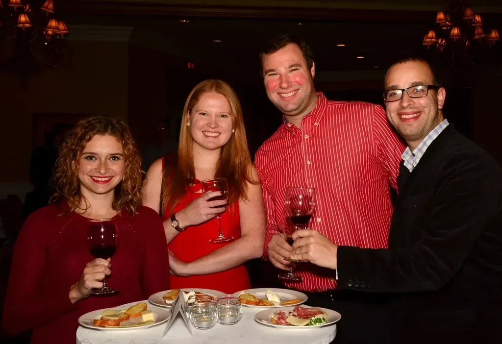 Four people at a gathering, holding wine glasses, smiling. Red and dark clothing, food on table.