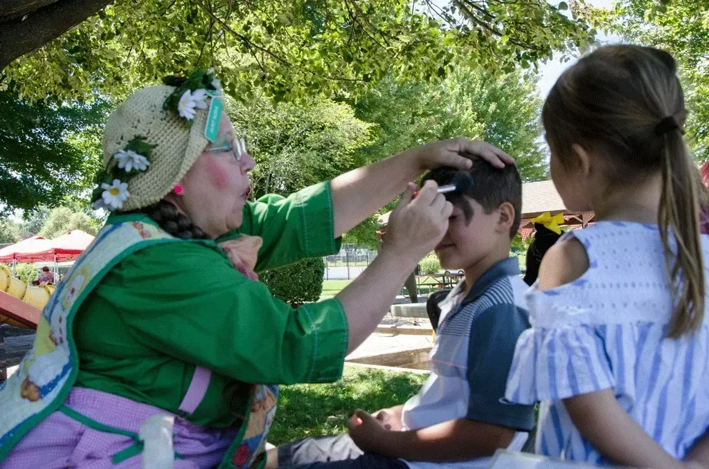 Person in costume paints boy's face outdoors, a girl watches.