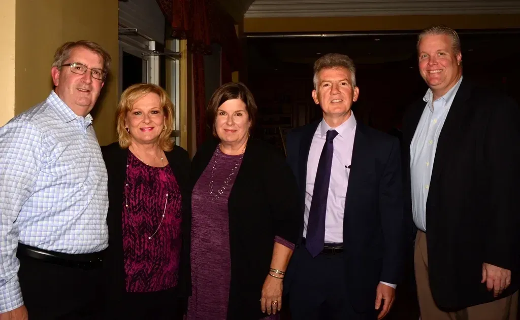 Five people posing together, smiling indoors. Men in suits, women in dresses, dark background.