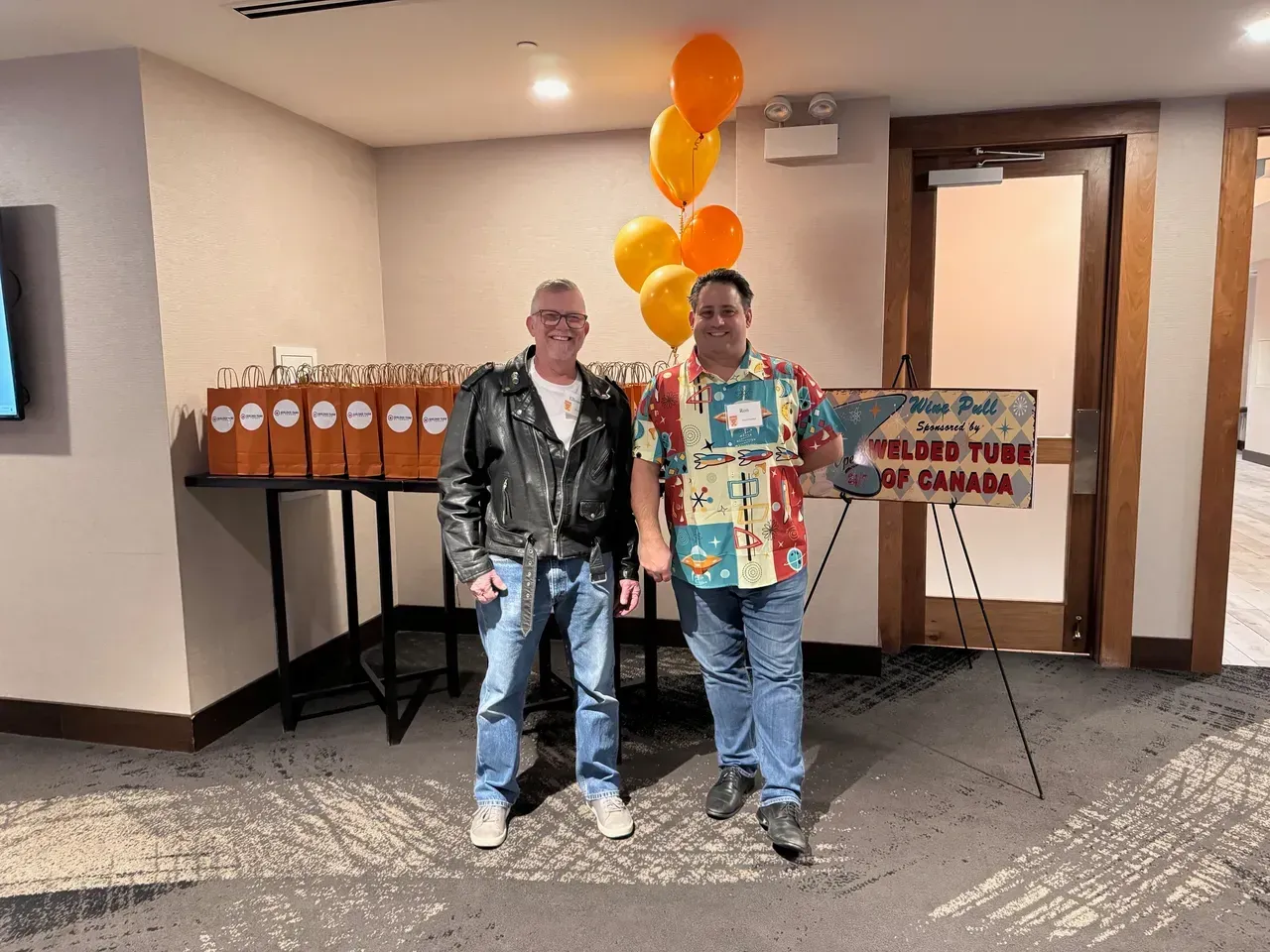Two men stand near a table with drinks and balloons. One wears a leather jacket, the other a patterned shirt.