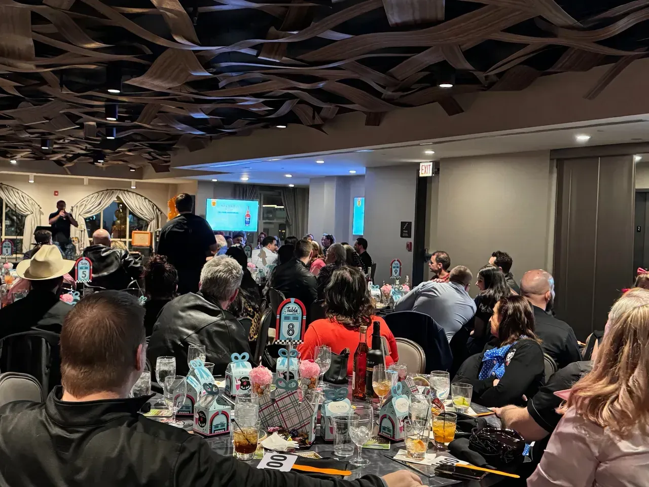 Large group at an event in a decorated room with people at tables facing a screen.