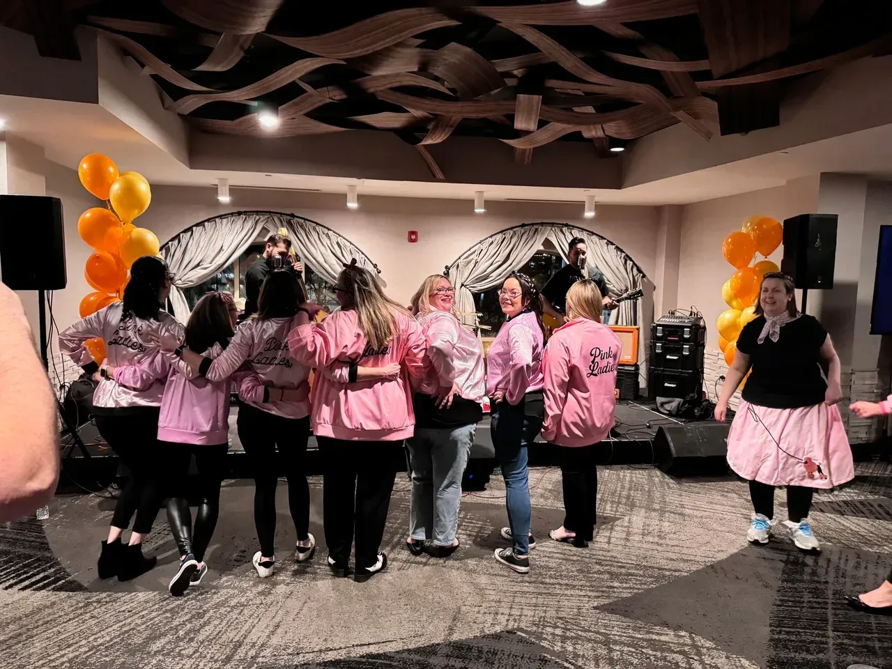 People in pink jackets and skirts dancing at a 50s-themed event with a band playing on a stage.