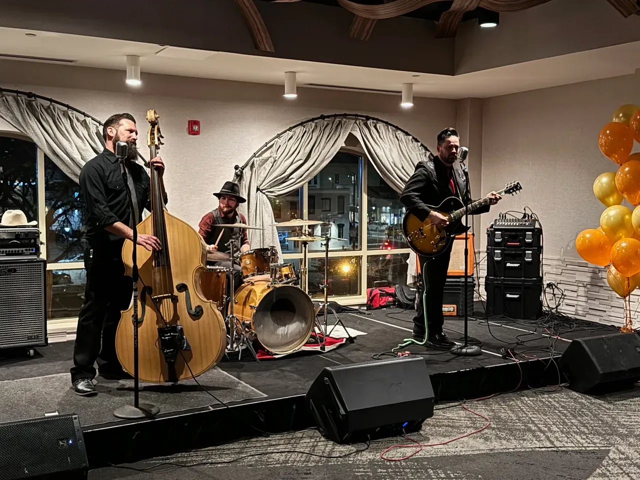 A band playing on stage: upright bass, guitar, drums. Black clothing, stage, and equipment. Balloons in the background.