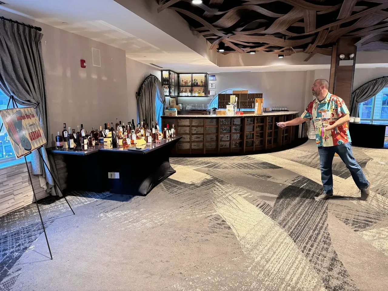 Man in Hawaiian shirt gestures toward wine bottles on a table near a bar in a dimly lit room.