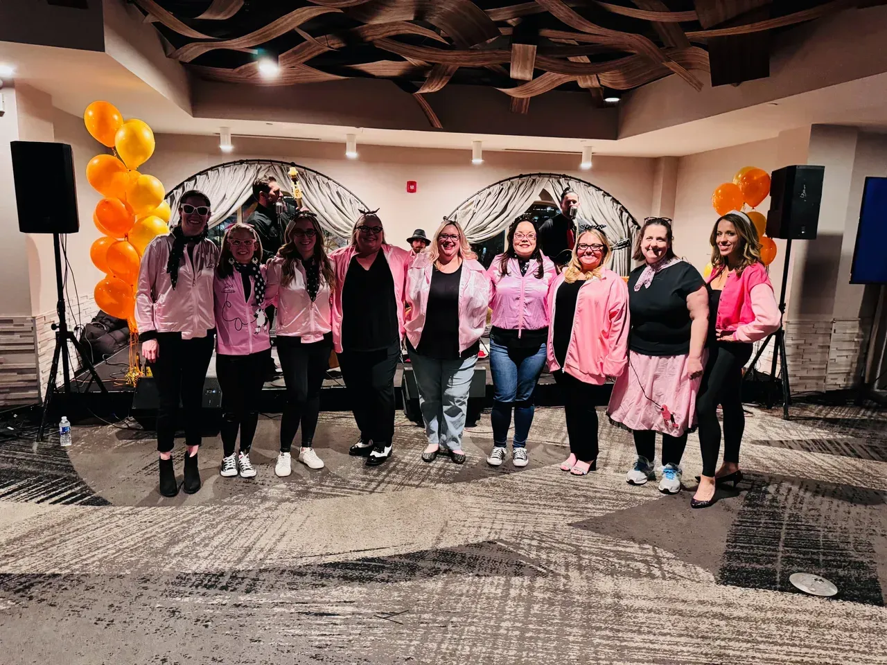 Group of women in pink jackets and outfits, posing indoors. Balloons in the background.