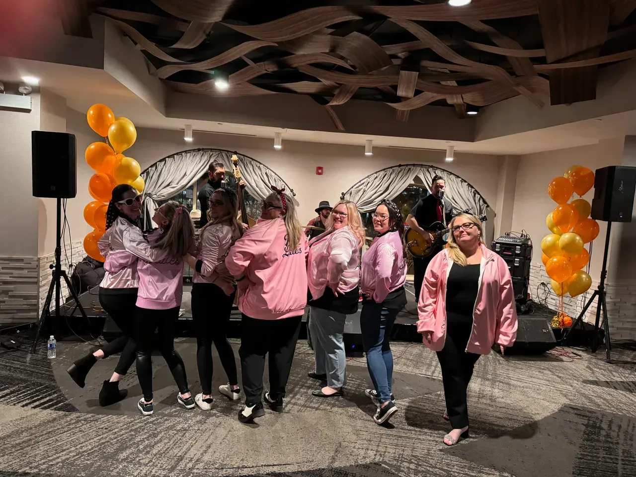 Group of women in pink jackets pose with a band in front of orange balloons.