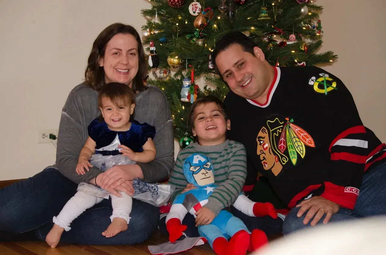 Family of four smiling in front of a Christmas tree; two children sit on parents' laps.