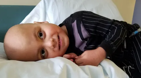 Child with a shaved head smiles, lying on a bed in what appears to be a hospital.