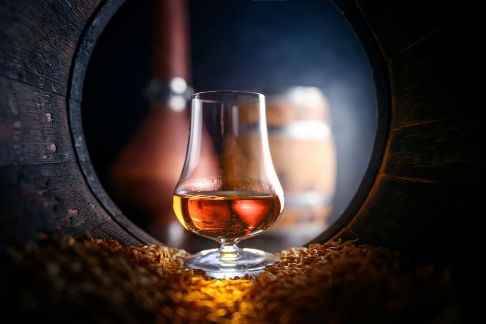 Glass of amber liquor inside a wooden barrel with blurred barrels in the background.