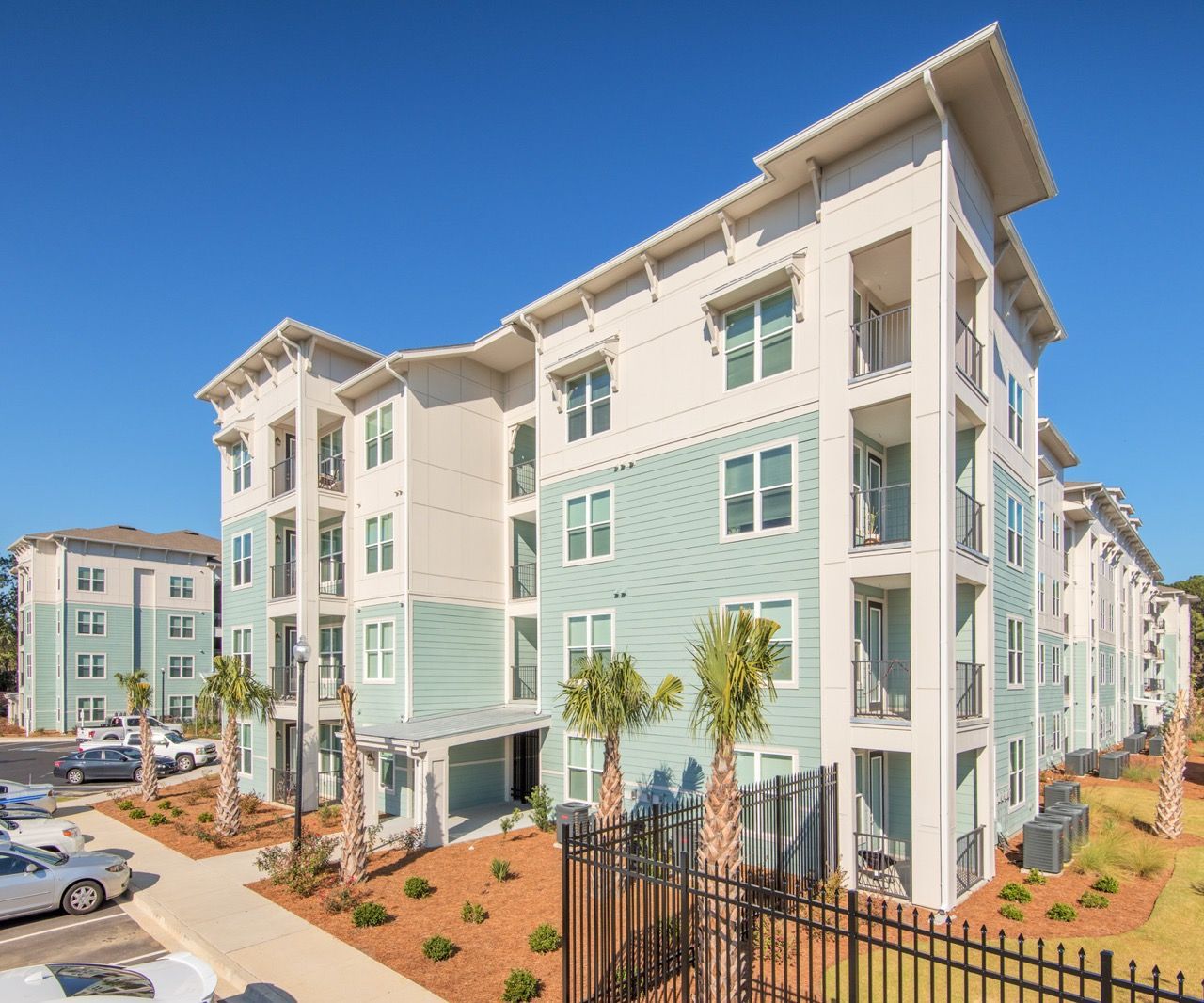 Exterior view of a modern apartment building with balconies and palm trees against a blue sky.