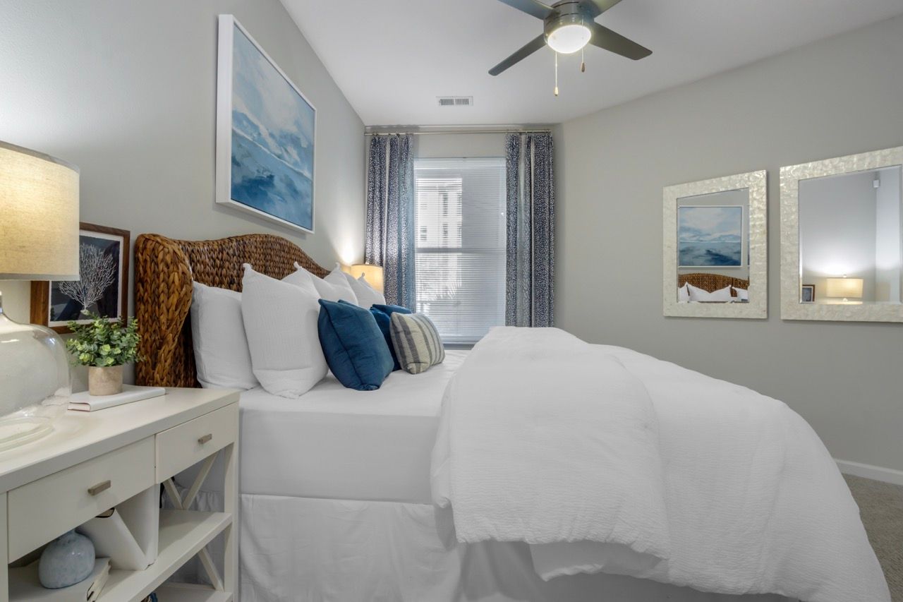 Bright modern apartment bedroom with white bedding, wicker headboard, blue accent pillows, and framed mirrors.