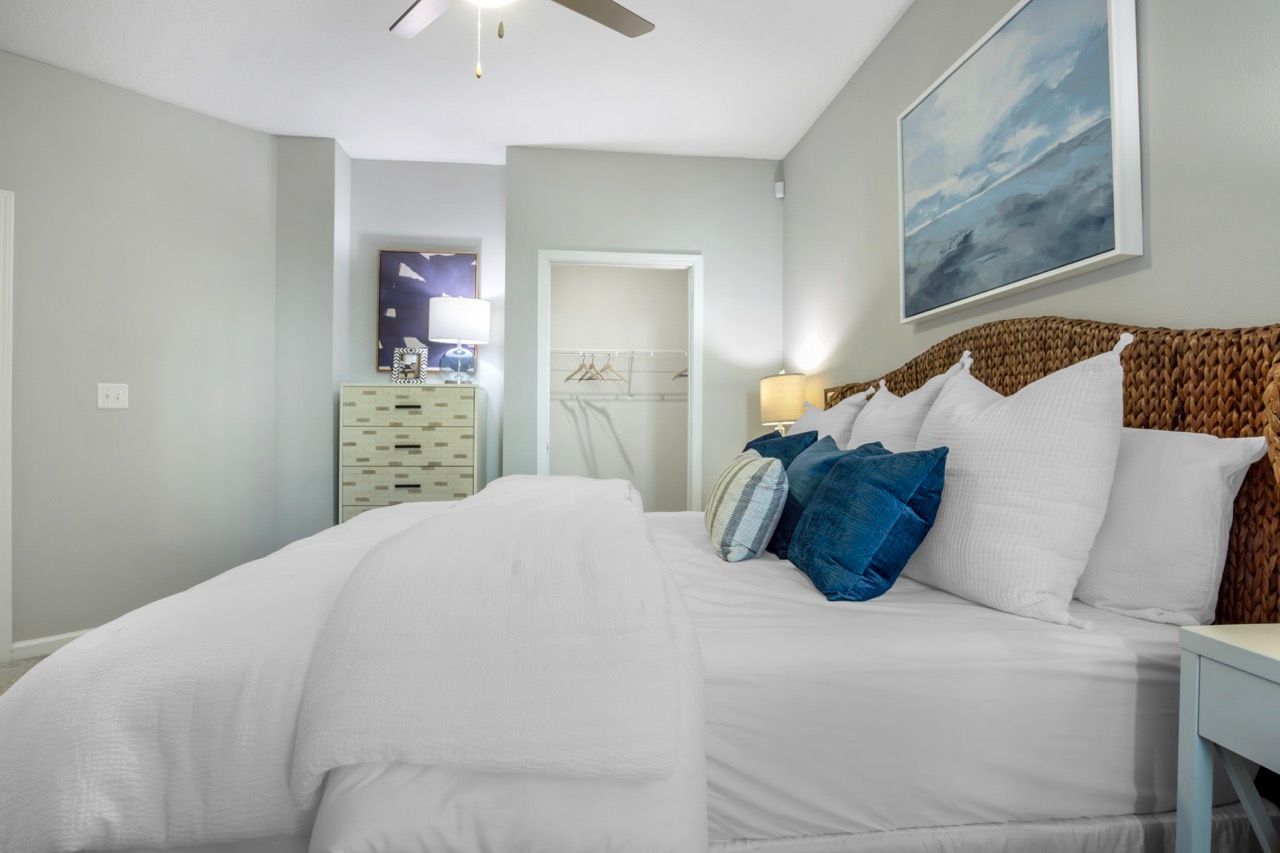 Bedroom in an apartment with a large white bed, wicker headboard, dresser, and open closet.