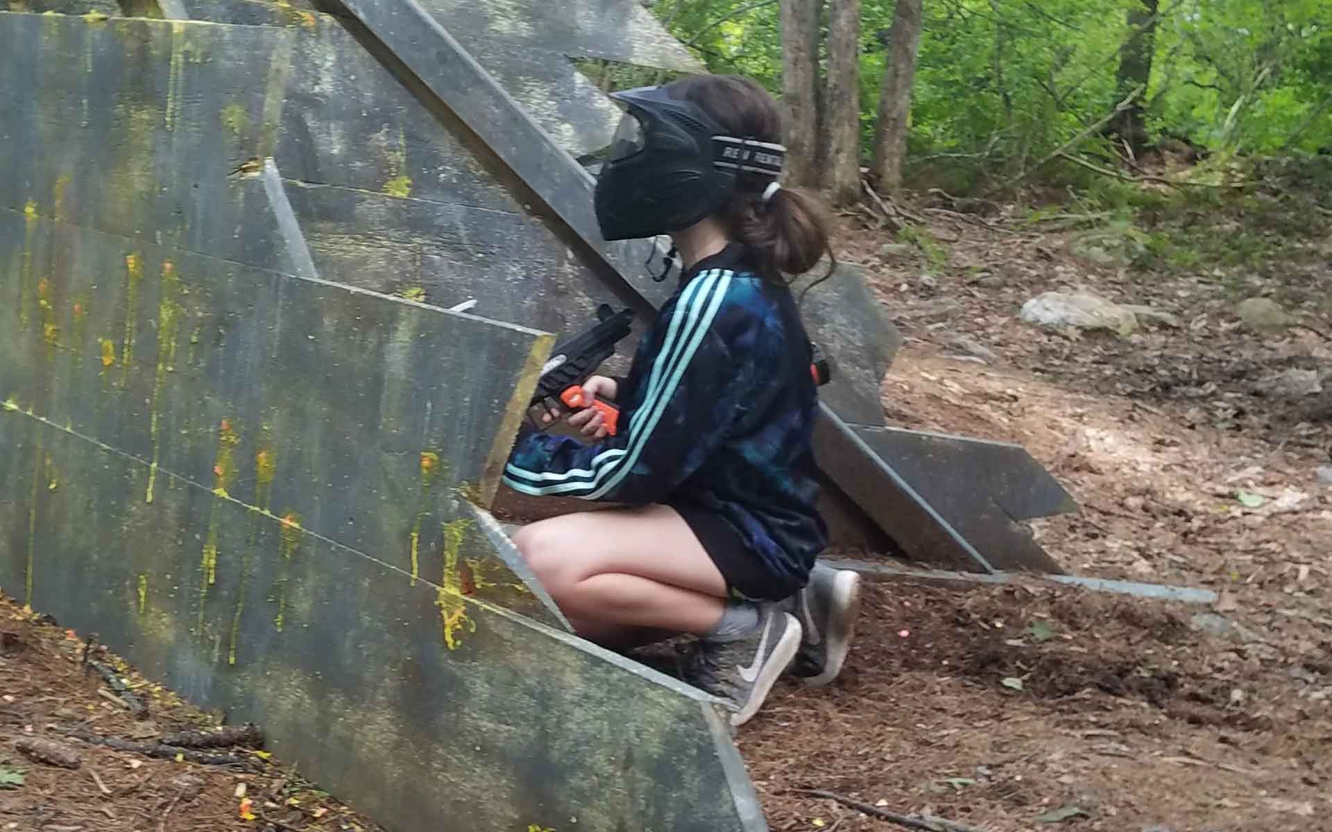 Person in paintball gear crouches behind barrier in wooded area.