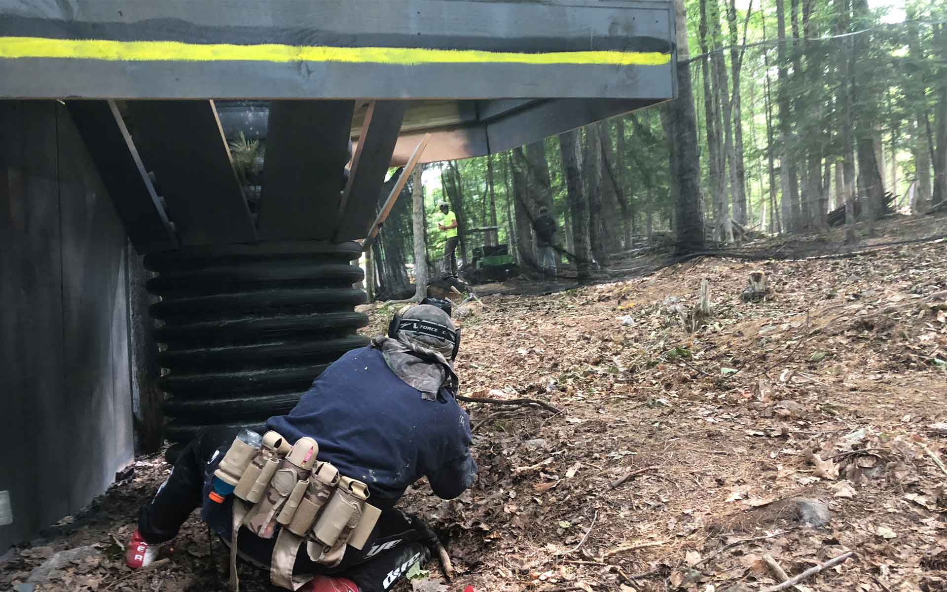 A paintball player crouches aiming, under a wooden structure. Forest setting.