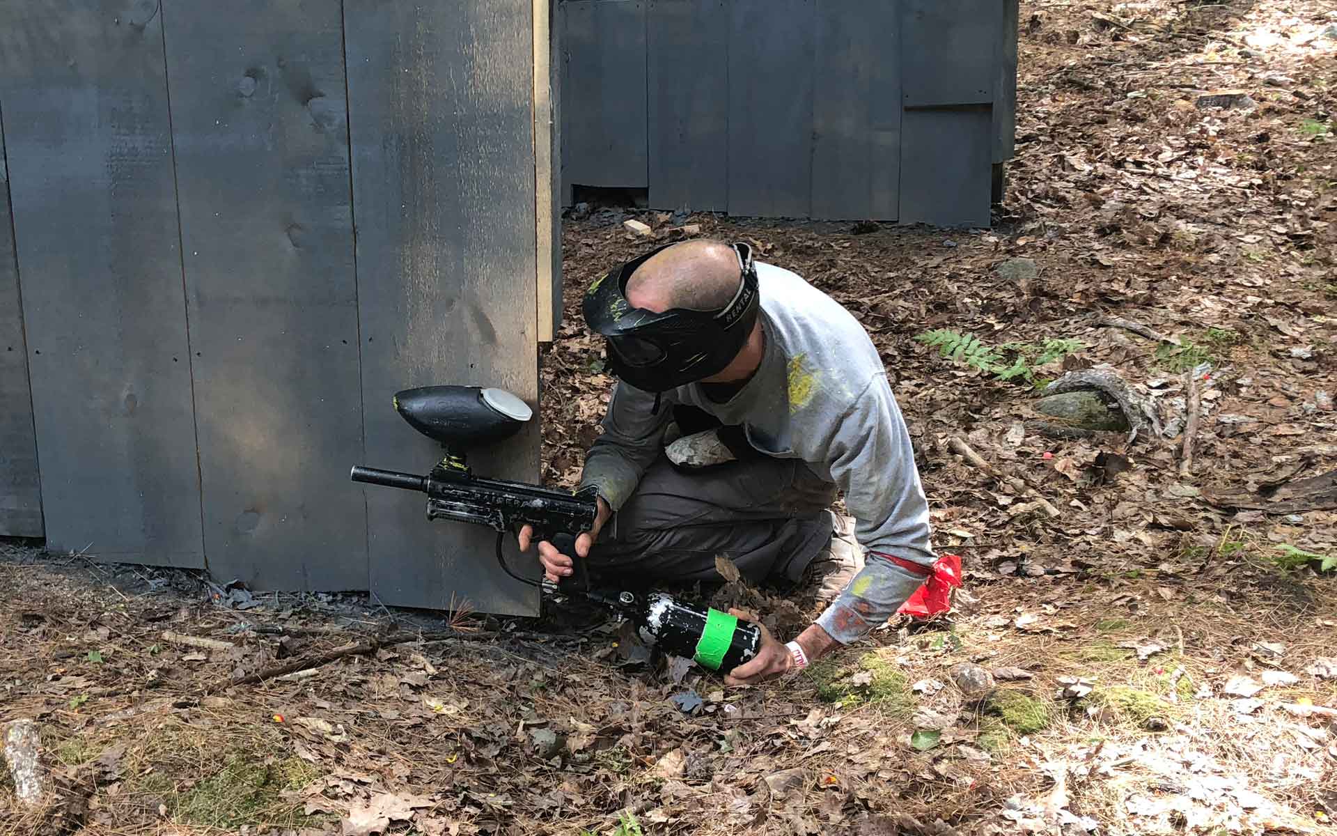 Man kneeling in paintball gear, reloading a gun, by a bunker in a wooded area.