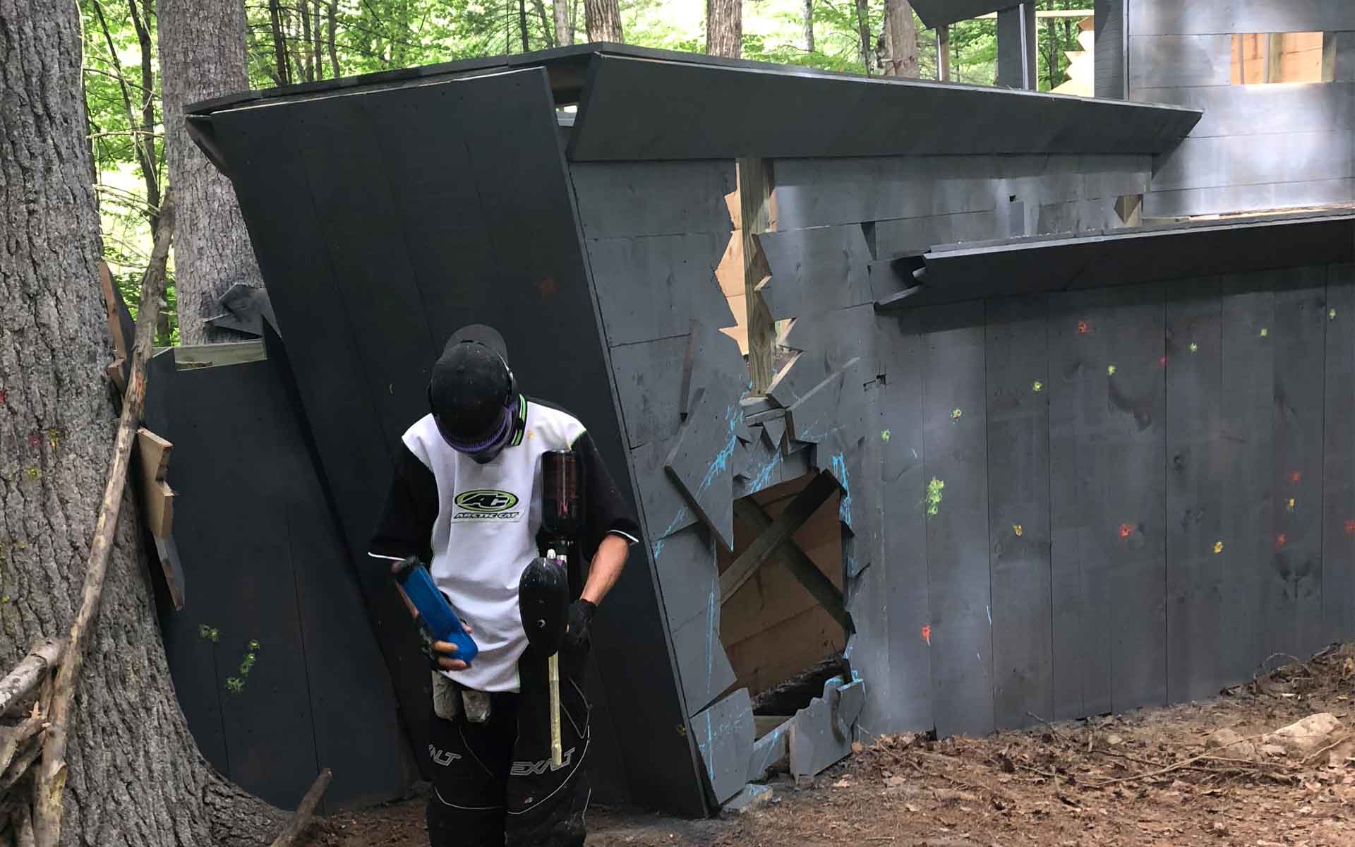 Person in paintball gear stands next to a dilapidated wooden structure with holes and splatters.