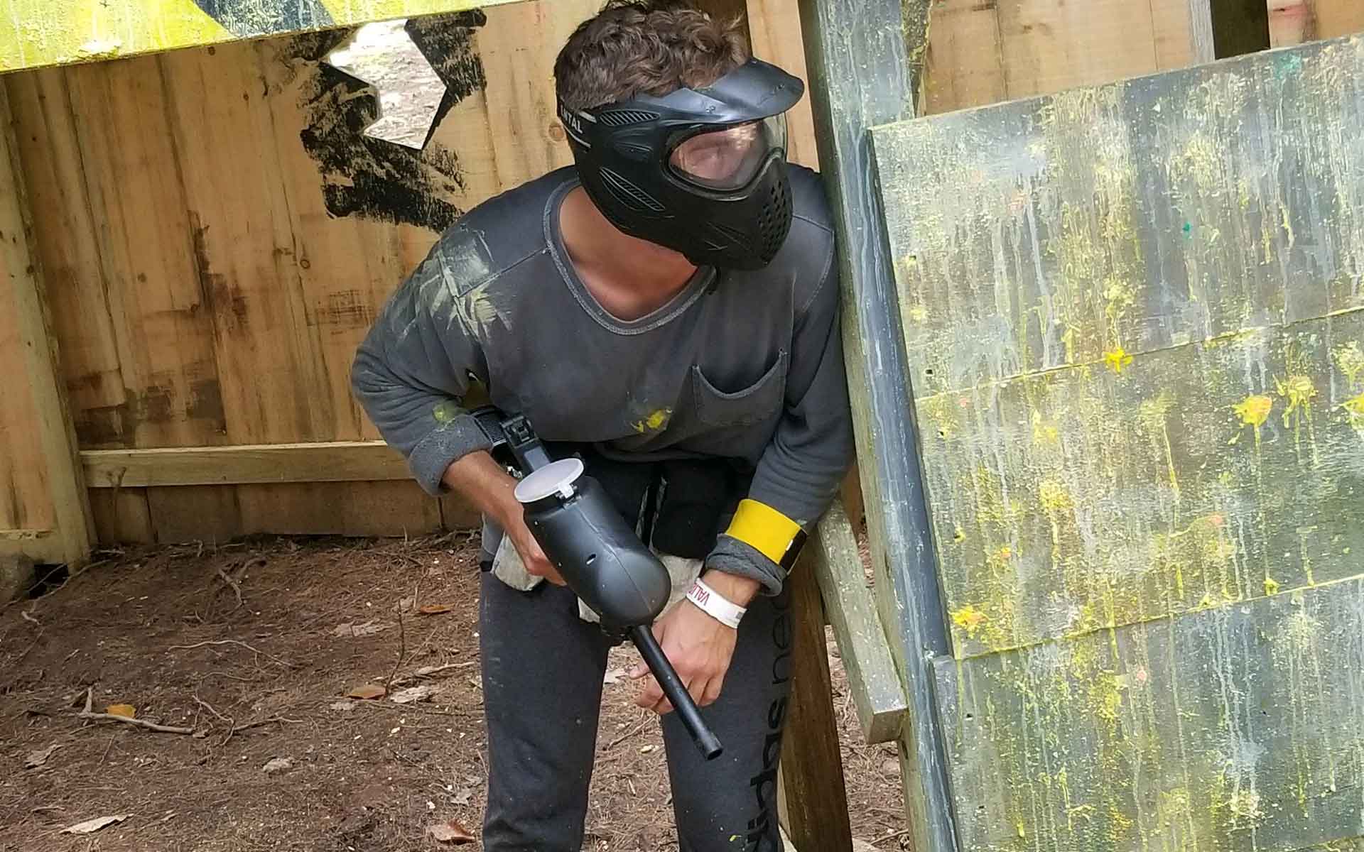 Person in black paintball gear, mask, holding a paintball gun, peeking around a wooden shelter.