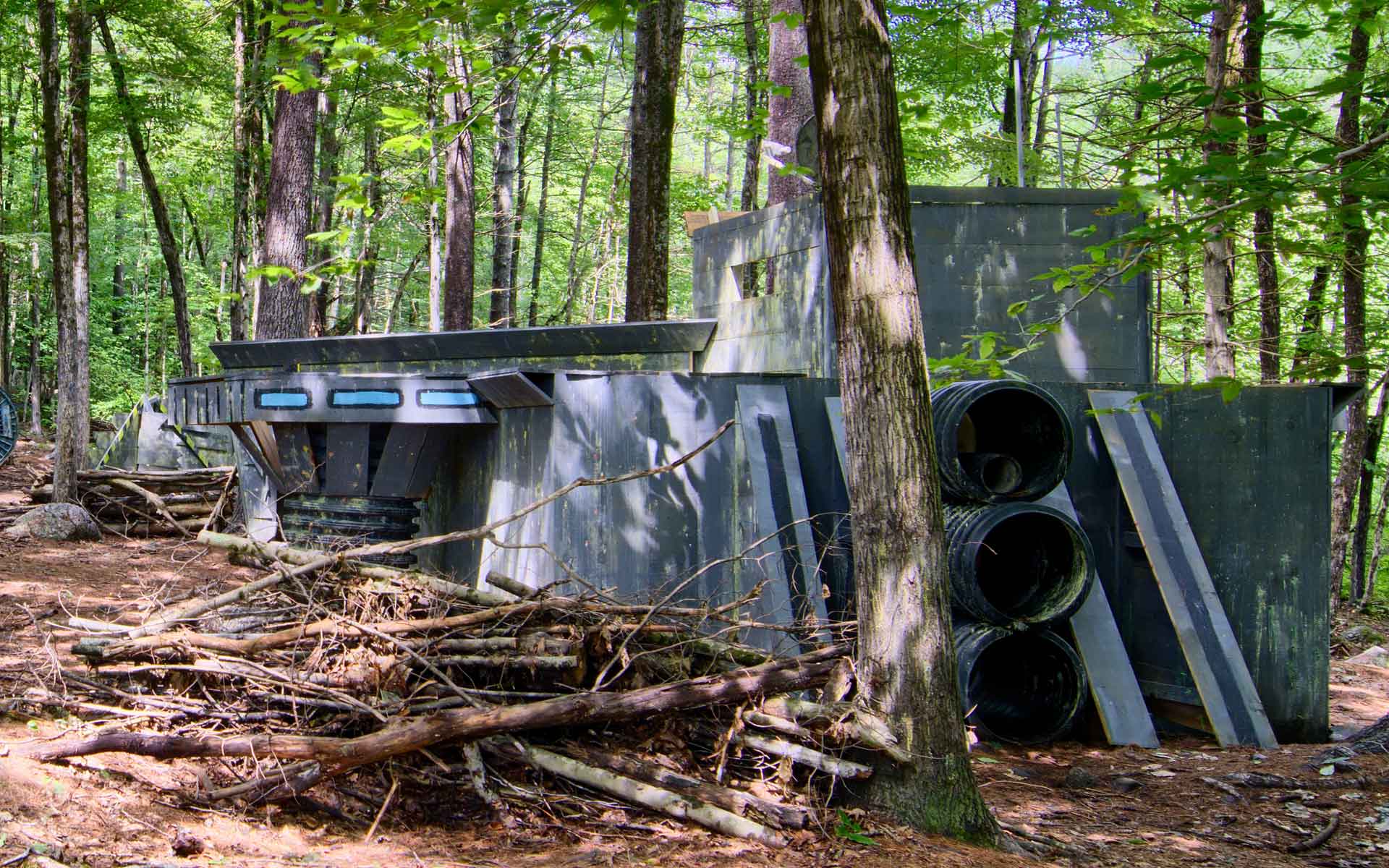 A paintball course structure in a wooded area, featuring grey barriers, tires, and blue accents.