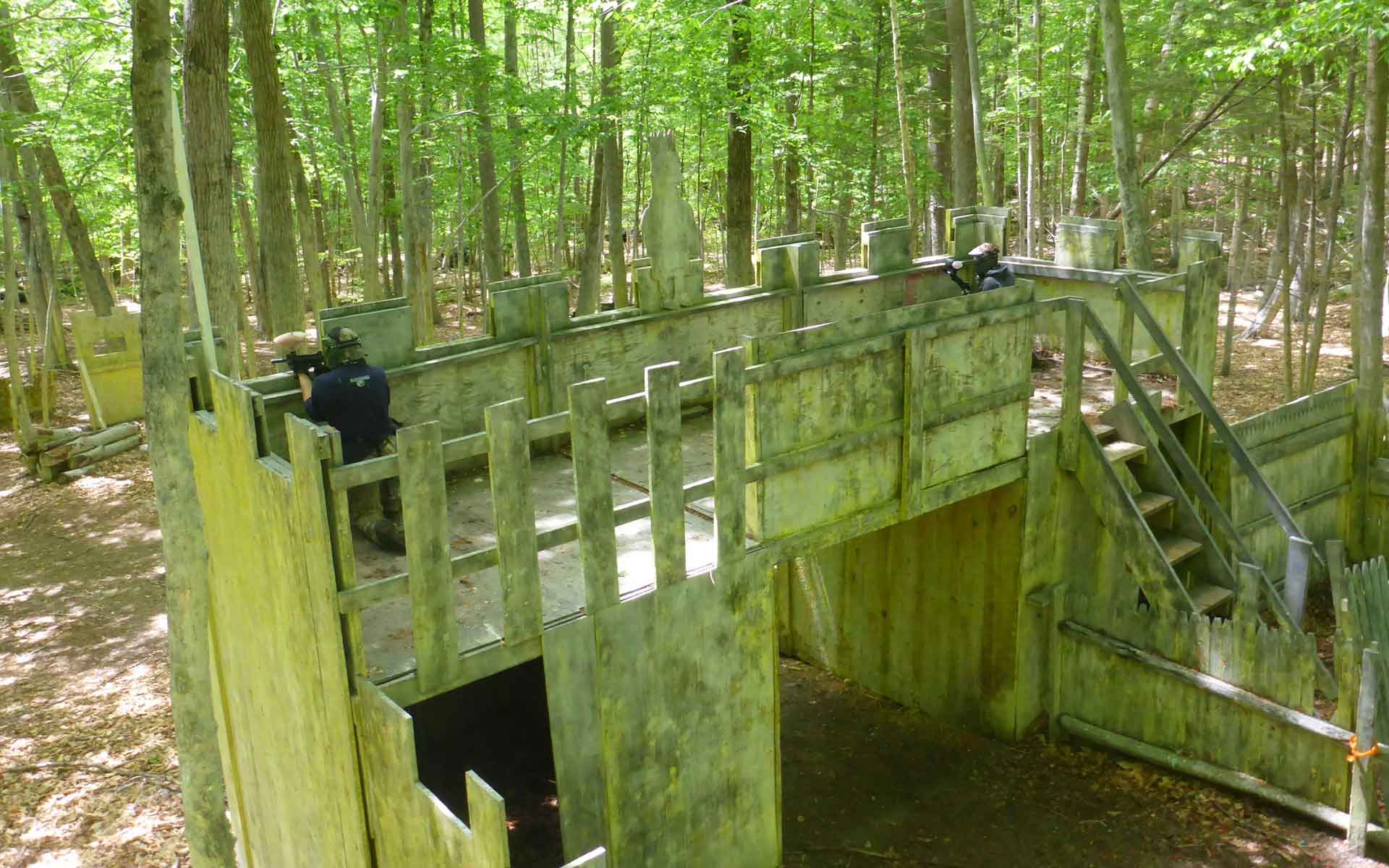 Paintball players on a wooden structure in a wooded area.